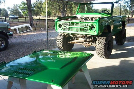 Green Ford Bronco with hood off, sitting outside next to a truck and a camper shell