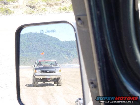 Dark Ford F-150 truck driving on a sandy beach, seen in a side mirror. Orange flag on antenna