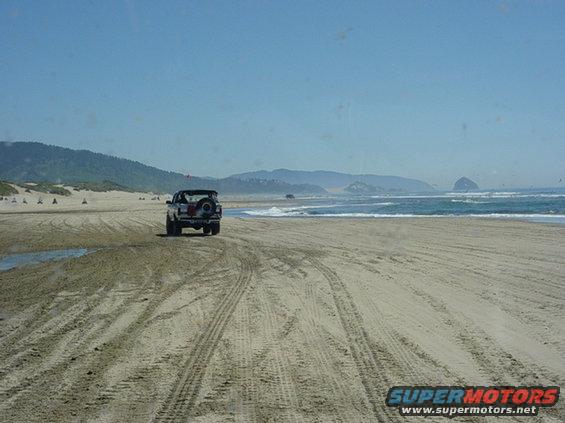 1995 Jeep Wrangler driving on a sandy beach with ocean and hills in background