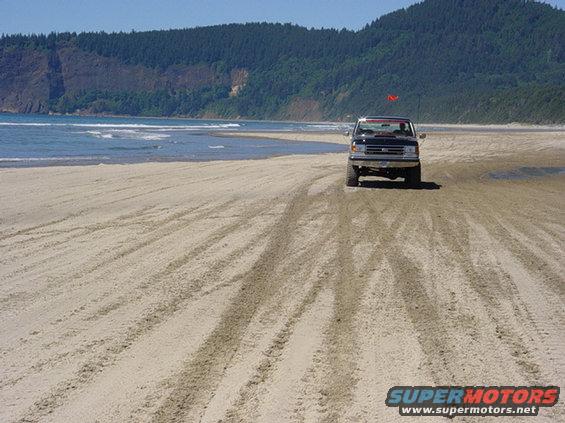 Black pickup truck driving on a sandy beach with ocean and hills in background