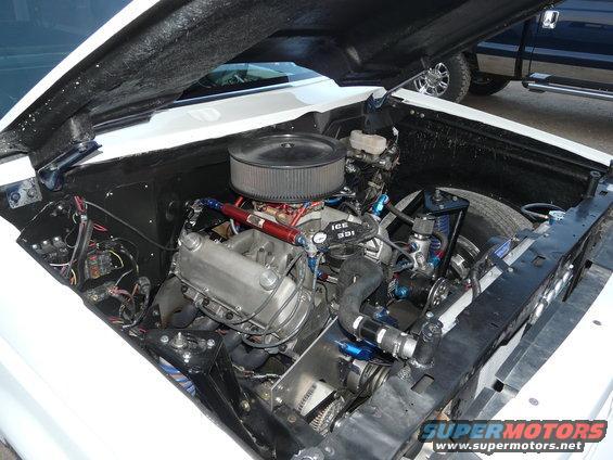 White vehicle's engine bay with a V8 engine, air filter, and blue hoses. A dark blue truck is visible in the background