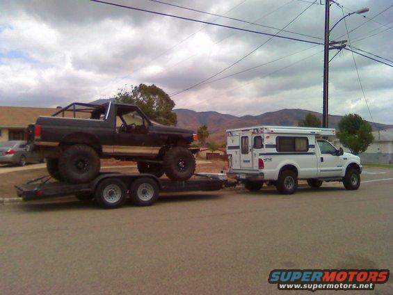 Black Ford Bronco on a trailer, towed by a white Ford F-250 with a camper shell, side view