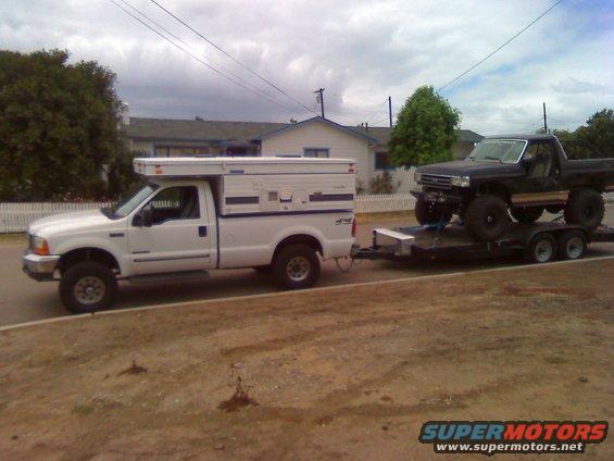 White pickup truck with camper towing a black pickup truck on a trailer