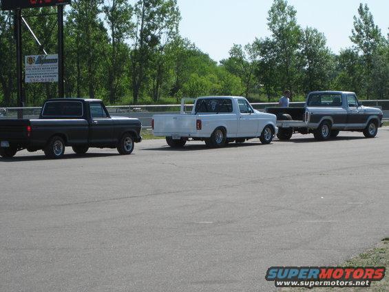 Three pickup trucks lined up on a paved surface, possibly a track. One is black, one is white, and one is two-tone grey and silver