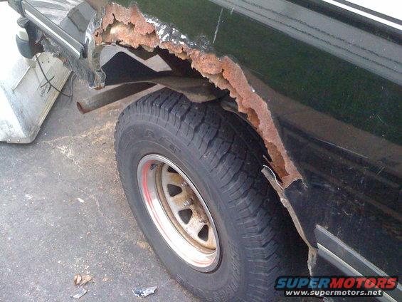 Rear wheel well of a dark vehicle with significant rust damage. A portion of a silver object is visible to the left