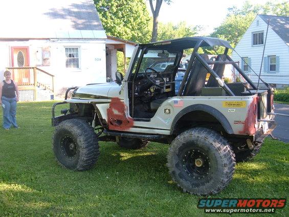 1976 Jeep CJ5 with large off-road tires, roll cage, and custom paint patches. A woman stands to the left