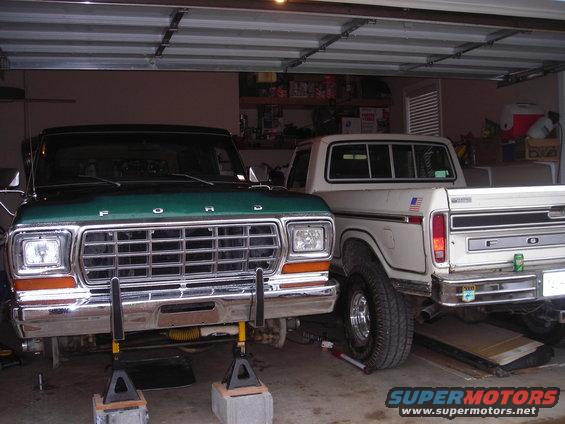 Green Ford truck and white Ford truck parked in a garage, both on jack stands