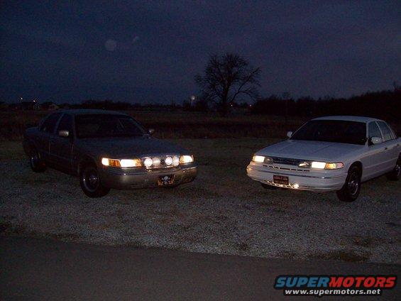 Two sedans, likely Ford Crown Victorias, parked outside at night. One is silver with extra lights on the grille, the other is white