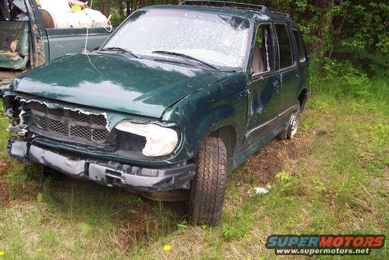 Damaged green SUV with front-end damage, missing headlight, and cracked windshield. Another vehicle is partially visible to the left
