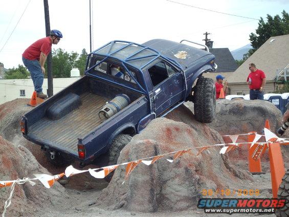 Blue pickup truck with a roll cage going over rocks. A man in a red shirt watches