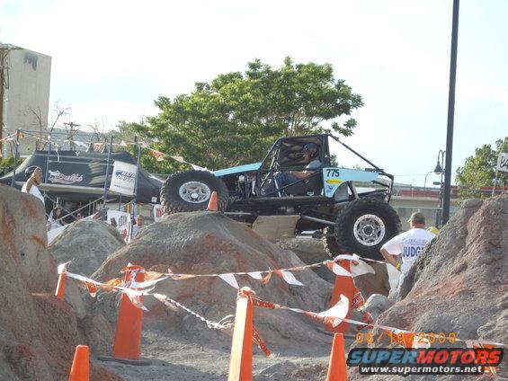 Off-road buggy with number 775 on the side climbing over rocks, with a judge watching