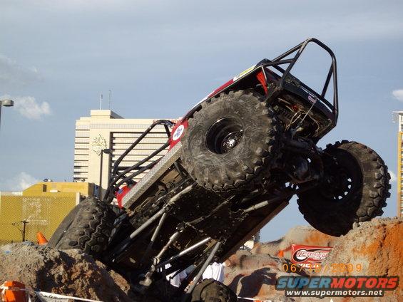 Off-road buggy with large tires climbing over rocks, with buildings in the background