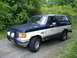 1988 Ford Bronco - A two-tone blue and white Ford Bronco with visible rust and aftermarket lights sits on a gravel road surrounded by greenery.