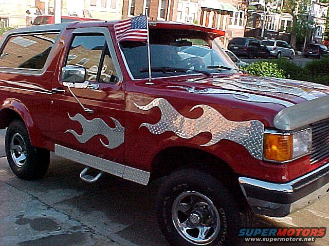 Red 1990 Ford Bronco with diamond plate flame decals and an American flag on the windshield