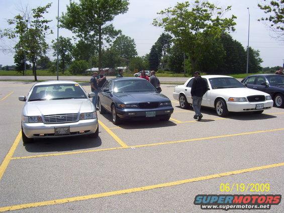 Three Ford Crown Victorias parked in a lot. One is silver, one is blue, and one is white. People are standing around them