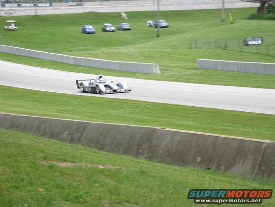 prototype race car on a track, with other cars parked on grass in background