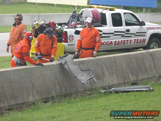 Damaged car part on grass near concrete barrier with SCCA Safety Team members and a white truck
