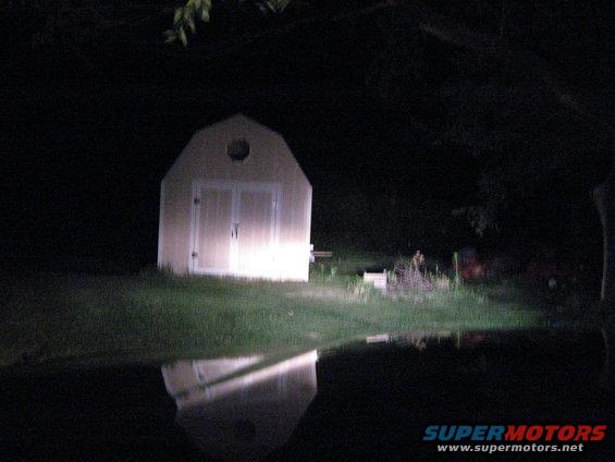 Shed with lights on, reflected in water at night. Supermotors.net logo in corner