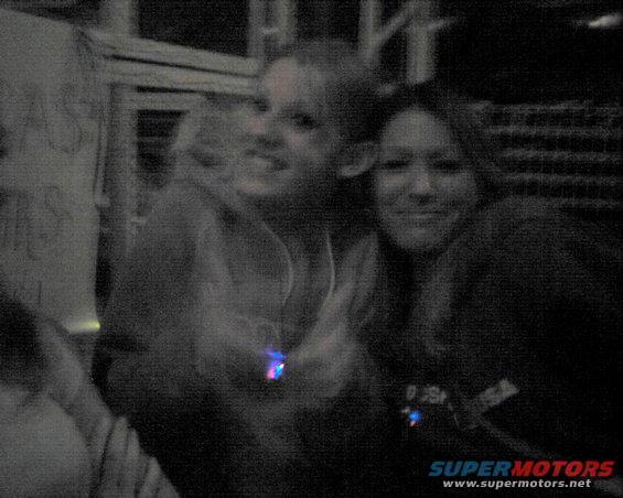 Two smiling people in a dimly lit indoor setting, with a "SUPERMOTORS" watermark at the bottom right