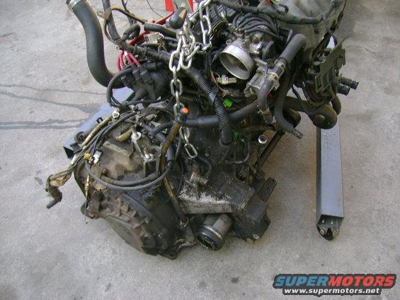 Engine assembly with transmission attached, covered in grease and grime, suspended by chains on an engine stand