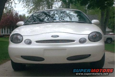 White 1997 Ford Taurus front view, grille and headlights visible, some trees and a white pickup truck in the background