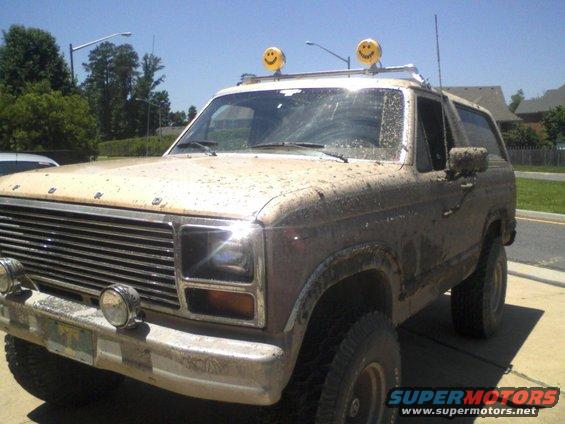 Mud-covered tan Ford Bronco with smiley face lights on roof rack, front 3/4 view