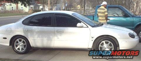 White sedan, side view, with tinted windows and alloy wheels. A teal pickup truck is partially visible to the right