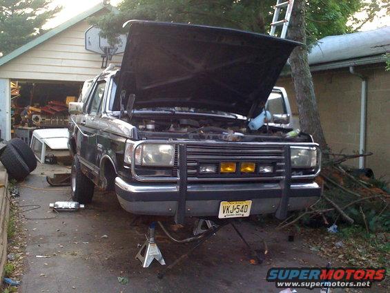 1987 Ford Bronco front view, hood open, on jack stands, in driveway with garage in background