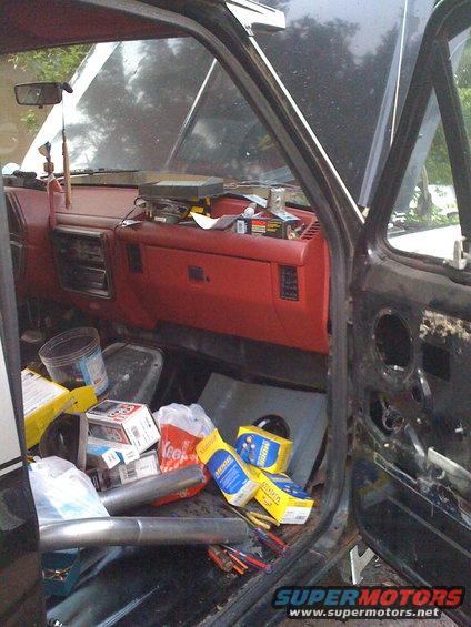 Interior of a vehicle with red dashboard, tools, and boxes of parts scattered on the floor. Hood is open
