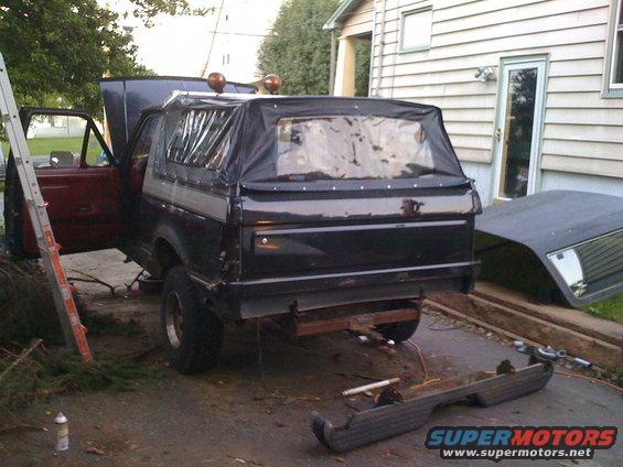 Black SUV with a soft top, rear view, in a driveway with a detached bumper and ladder