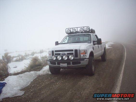 White Ford F-Series truck with roof rack and bumper lights parked on a dirt road in foggy weather