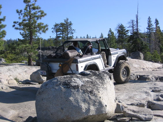 White Ford Bronco with off-road tires navigating rocky terrain, driver visible
