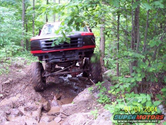 Red and black off-road truck with large tires navigating a muddy, rocky trail in a forest