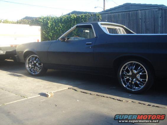 1973 Ford Ranchero with aftermarket chrome wheels, matte black paint, parked next to a trailer