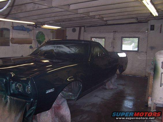 Dark green vehicle body, partially covered, on stands in a garage. Front fender and headlight visible