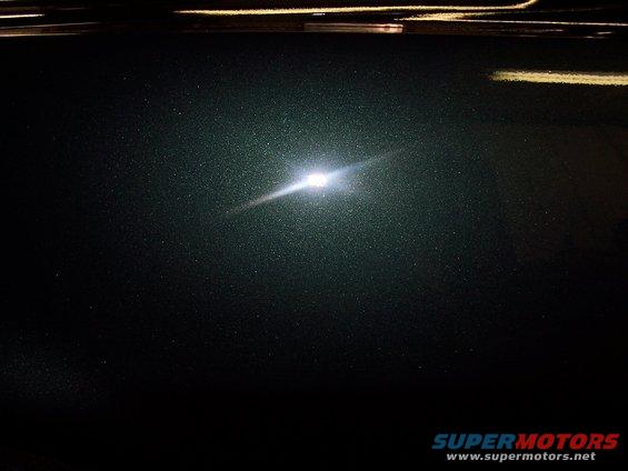 Close-up of dark green metallic paint with a bright light reflection. Supermotors.net logo visible