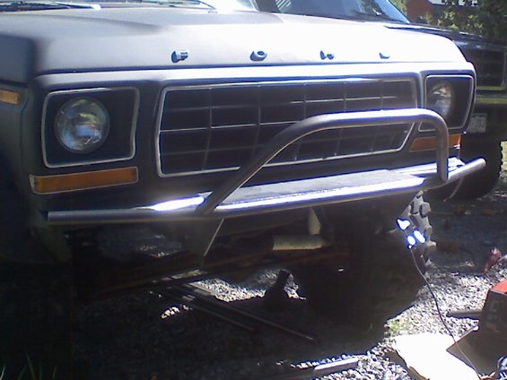 Front of a dark gray Ford truck with a custom bumper and grille. Another vehicle is visible behind it