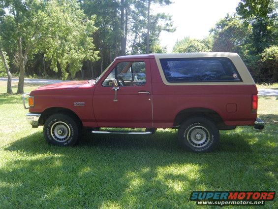 Side view of a red 1989 Ford Bronco with a tan removable top, parked on grass