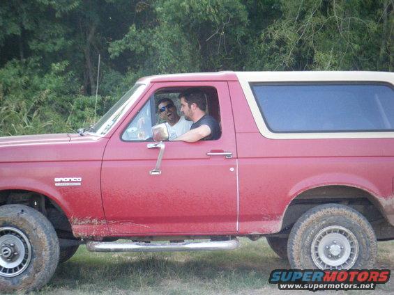 Red 1989 Ford Bronco side view with two men in the cab, mud on the lower body