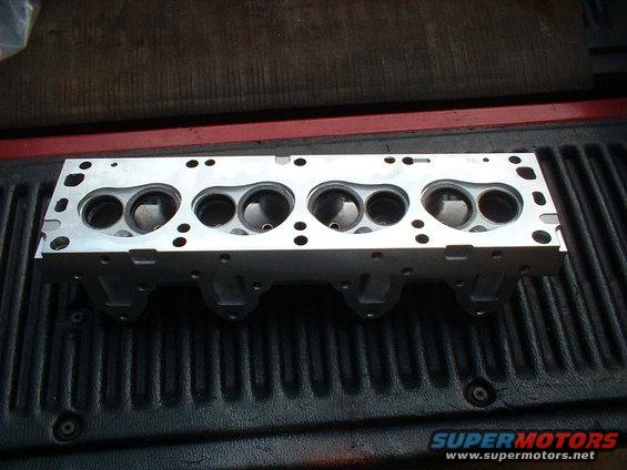 Aluminum engine cylinder head with six combustion chambers, resting on a black ribbed surface