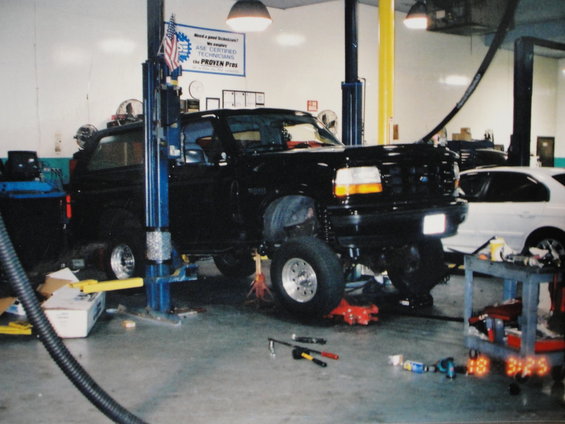 Black SUV on a lift in a garage, with a white car in the background. Tools are on the floor