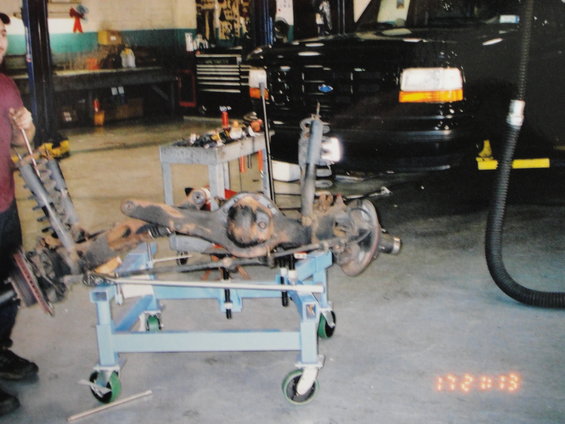Vehicle rear axle assembly with shocks and brakes on a blue cart in a garage. A black truck is visible in the background