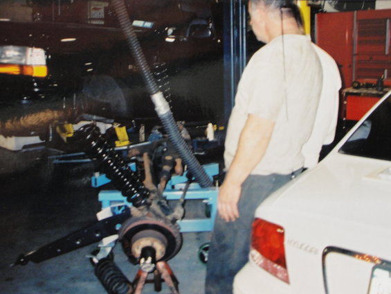 Mechanic working on front suspension of a vehicle in a garage. Another car is parked nearby
