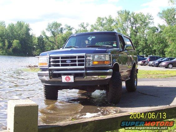 Blue Ford Bronco with a brush guard, partially in water next to a parking lot