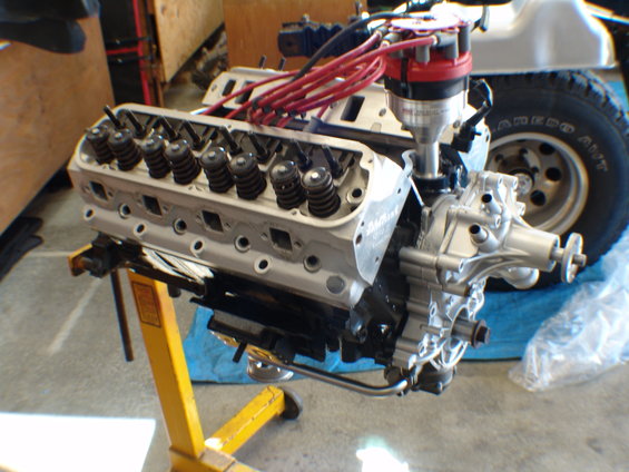 Engine block with valve covers, spark plug wires, and distributor cap. A tire and part of a vehicle are visible in the background