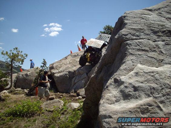 Off-road vehicle with exposed engine and roll cage climbing a large rock face, with people watching