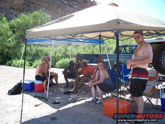 Group of people sitting under a canopy on a sandy area near trees and a river