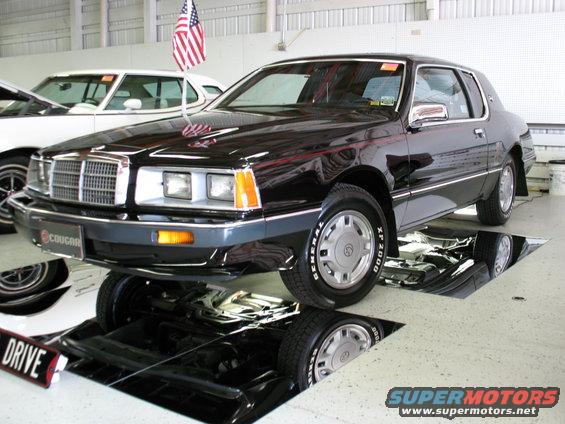 1985 Mercury Cougar with American flag on antenna, parked inside a garage with a pit