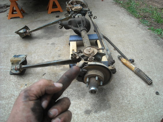 Front axle assembly with disc brakes and tie rods, next to a shock absorber on concrete. A dirty hand is in the foreground