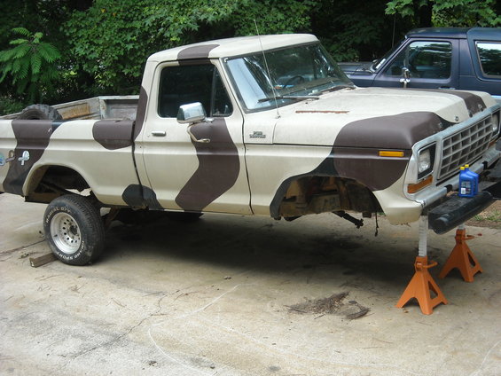 1979 Ford F-150 pickup truck with camouflage paint, missing front wheels, on jack stands. Another dark blue vehicle is parked behind it